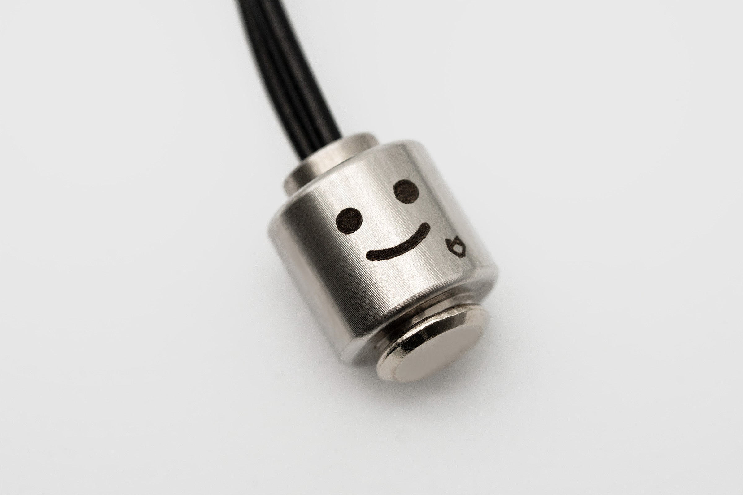 Close-up of Little Lupo Stainless Steel Pendant with a happy face expression, showing its brushed metal finish and tiny brand logo, designed to attach to dog leashes.