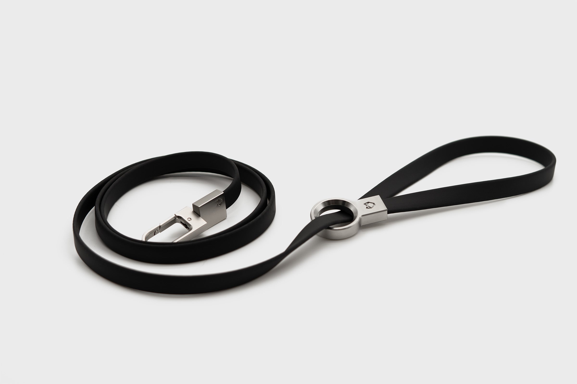 Little Lupo Leash L3 coiled on a white surface, highlighting its minimalist design, flexible black silicone-coated nylon webbing, and elegant stainless steel clip and handle ring.