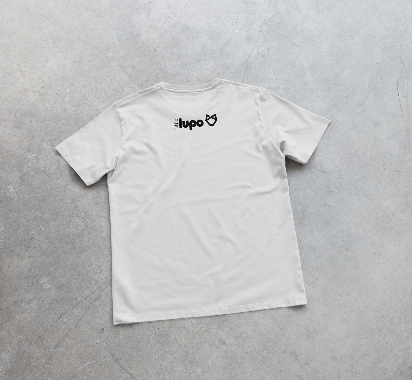Back view of a white Little Lupo t-shirt laid flat on a concrete surface to show the full design featuring the Little Lupo logo located at the top center. 