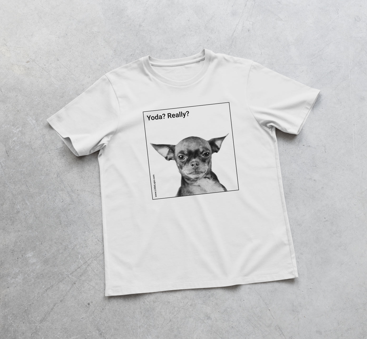 Top view of a white Little Lupo t-shirt laid flat on a concrete surface to show the full design featuring a black and white image of a dog resembling Yoda from Star Wars, with the text 'Yoda? Really?'.